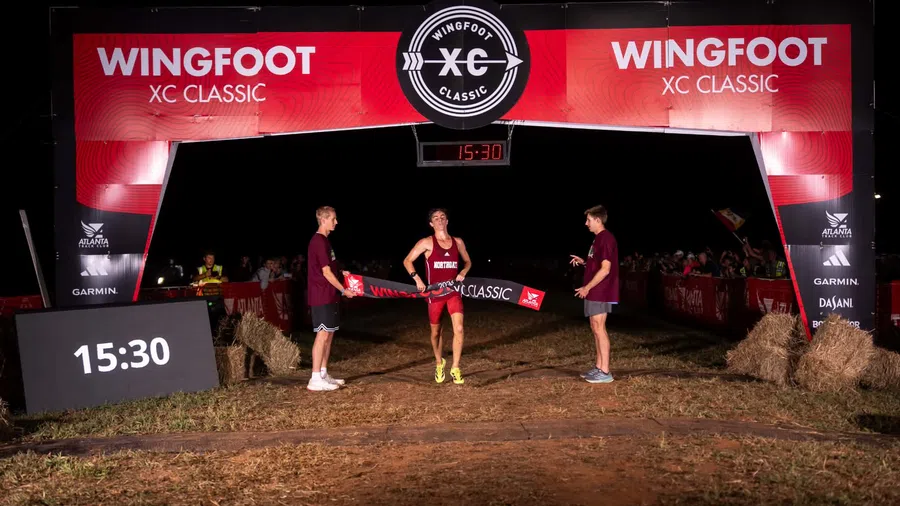 Runner crosses finish line at Wingfoot XC Classic race under illuminated event arch at night.