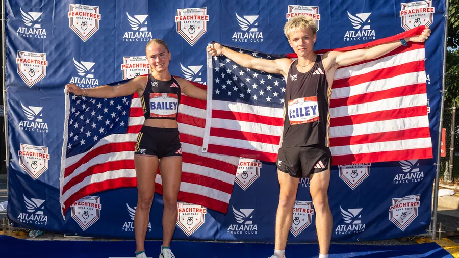 Two young athletes, Bailey and Riley, pose holding American flags at the Peachtree Road Race backdrop in Atlanta.