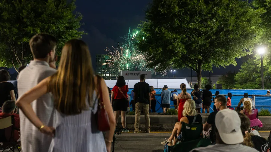 Crowd watching colorful fireworks display at night near Macy's store with trees and streetlights around.