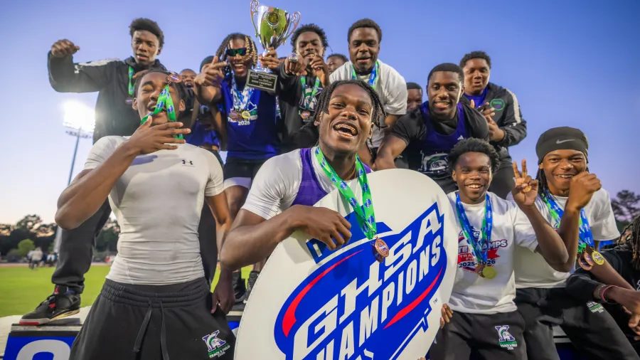 Excited football team celebrating GHSA championship win with trophy, medals, and sign at sunset on field.