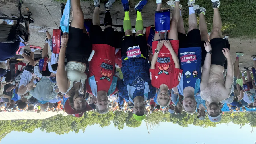 Group of six runners posing with race bibs and medals outdoors on a sunny day with crowd in background