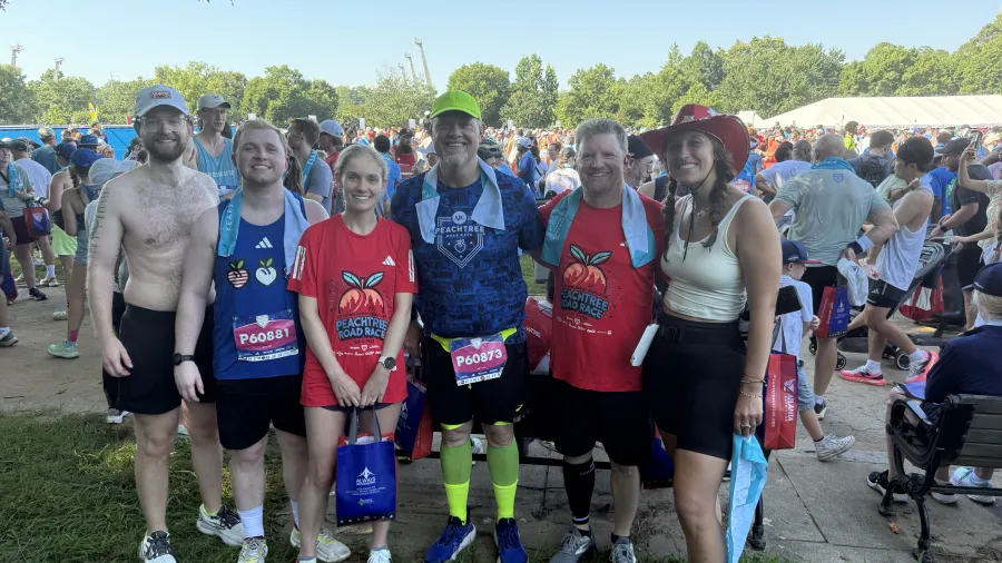 Group of six runners smiling at outdoor event with race bibs, medals, and crowd in background on sunny day