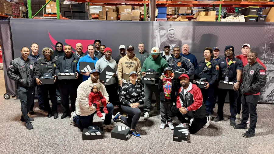Group of diverse people smiling and holding Adidas shoe boxes inside a warehouse with shelves and products.