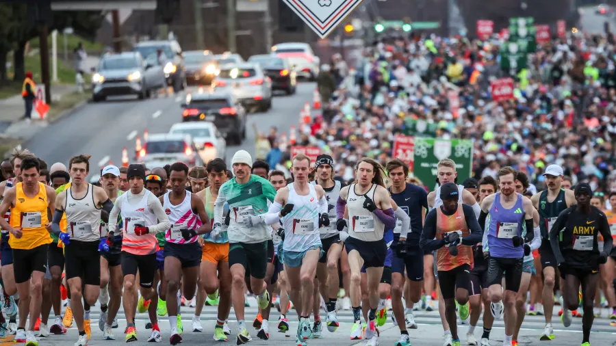 Runners start a marathon race under a green Marathon Weekend banner, with a large crowd behind them.