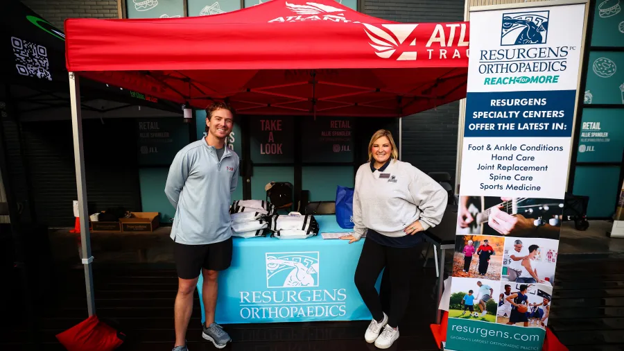 Two representatives at Resurgens Orthopaedics booth with red canopy and informational banner outdoors.