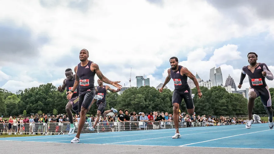 Sprinters racing on blue track in front of crowd with city skyline and cloudy sky background.