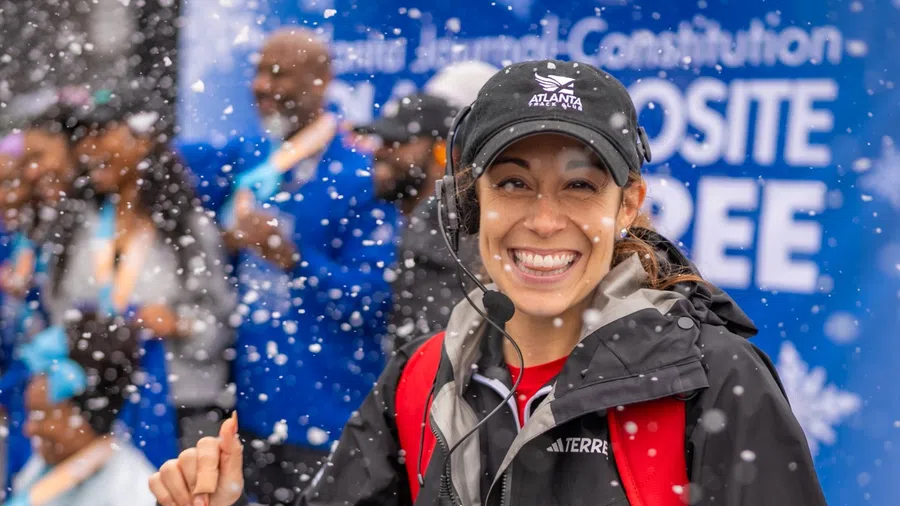Smiling woman in black jacket and cap with headset amidst falling snow at an outdoor event with blurred crowd background.