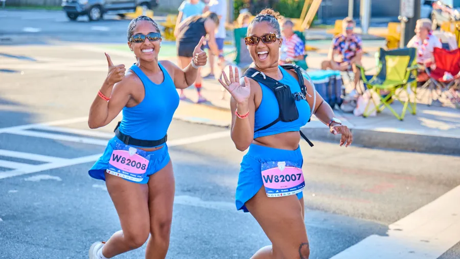 Two women wave and smile while running the Northside Hospital Peachtree Road Race