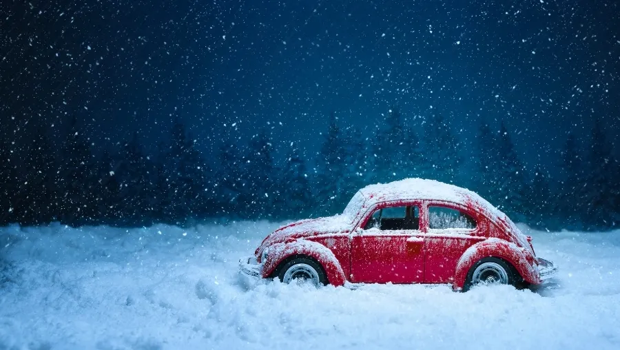 Red vintage car covered in snow driving through a snowy landscape under a dark blue winter sky.