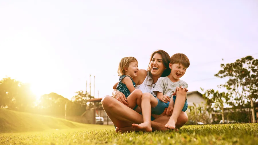 Smiling mother sitting on grass outdoors with two laughing children during sunset in a park setting