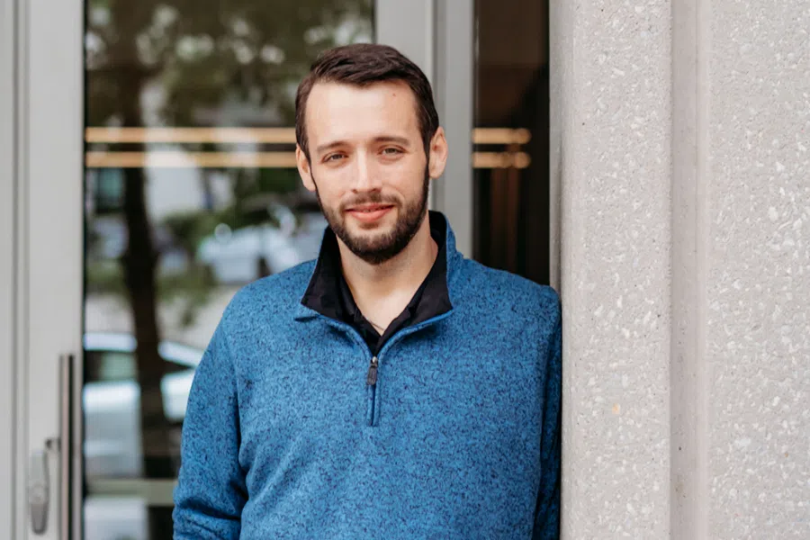 Smiling young man with beard wearing blue pullover standing outdoors near a building wall