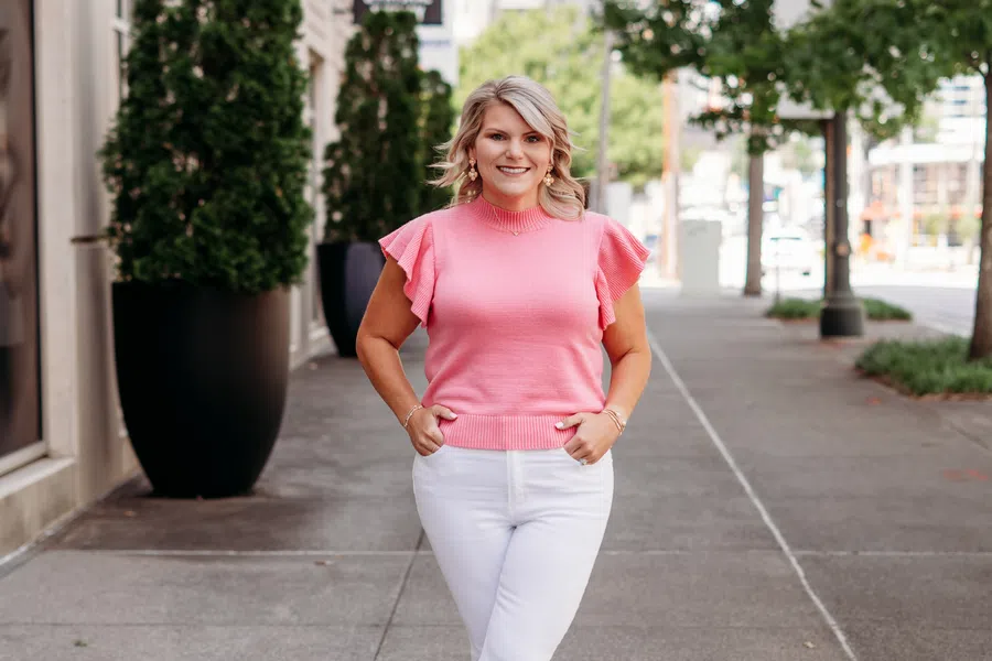Smiling woman in pink top and white pants standing on urban sidewalk with trees and buildings.