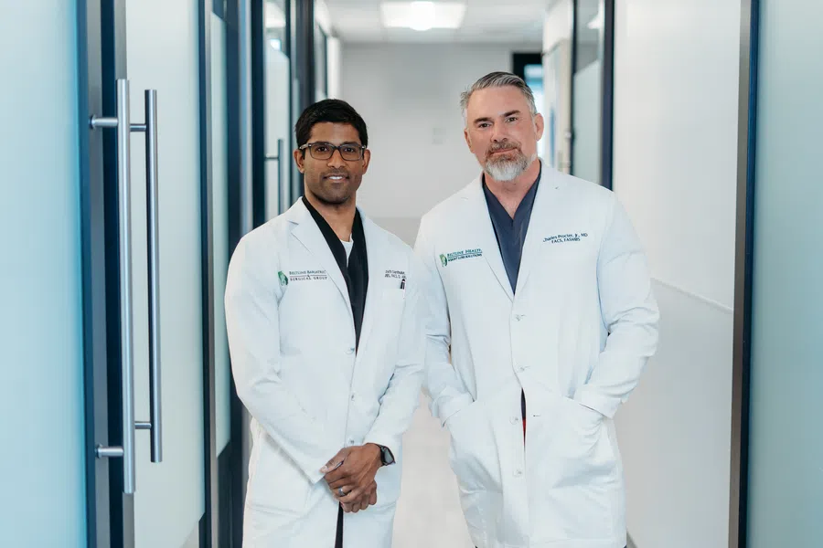 Two male doctors in white coats standing in a modern medical clinic hallway, smiling at the camera