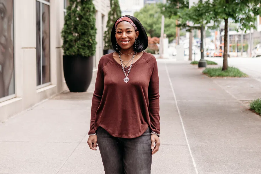 Smiling woman in casual brown top, black pants, and boots standing on urban sidewalk with trees and buildings.