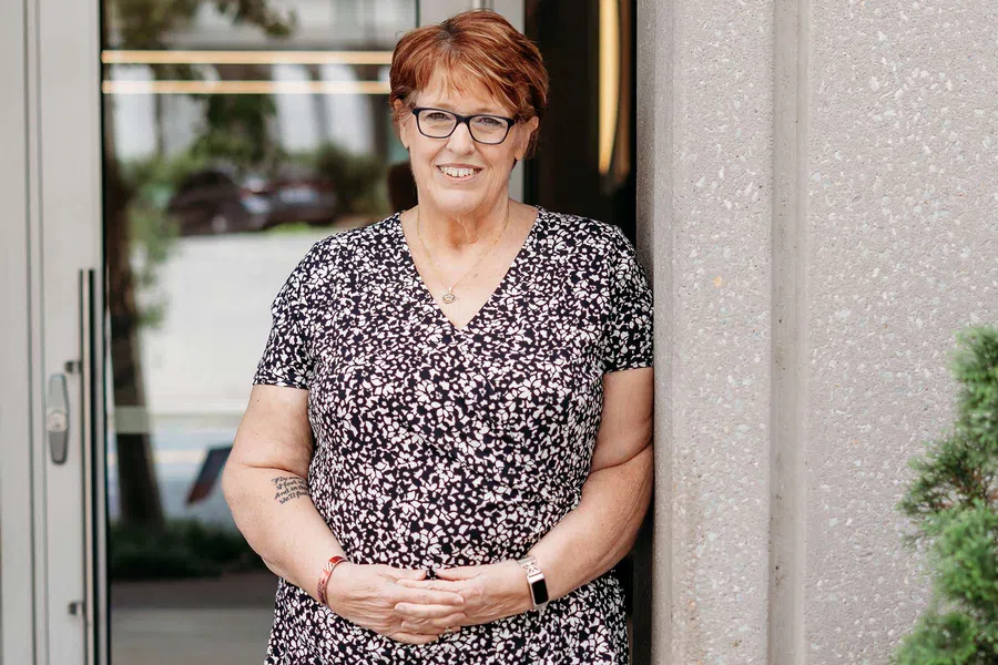 Smiling middle-aged woman with short red hair and glasses standing outdoors by a gray wall in floral dress.