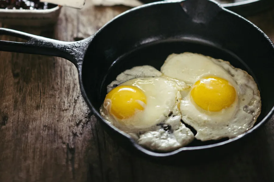 Two sunny-side-up eggs in a black cast iron skillet on a rustic wooden table with breakfast sides blurred.