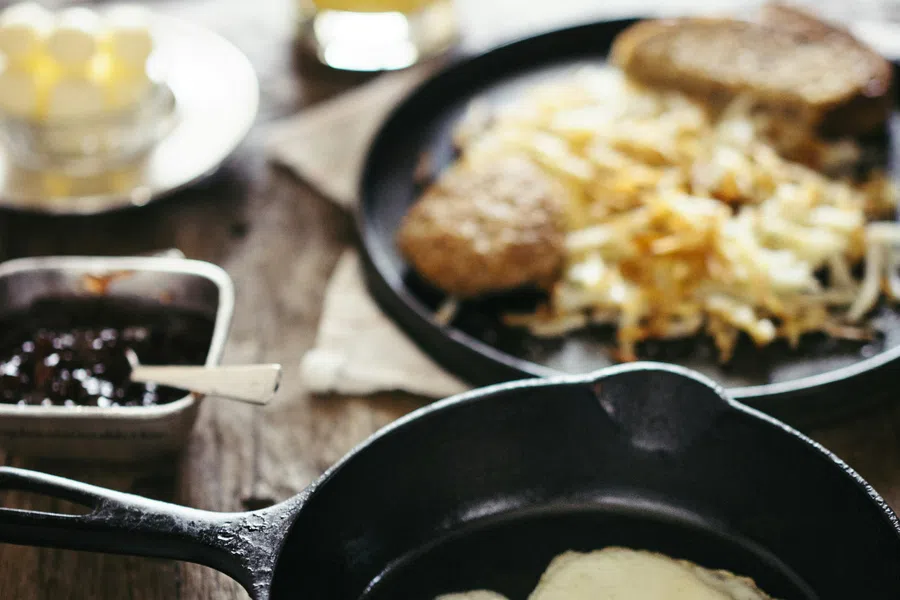 Two sunny-side-up eggs in a black cast iron skillet on a rustic wooden table with breakfast sides blurred.