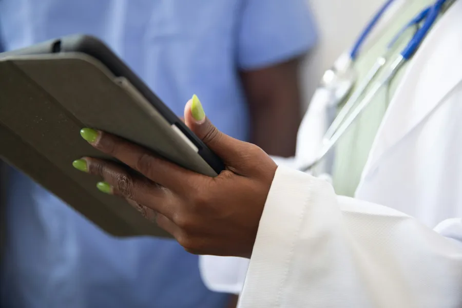 Close-up of a doctor holding a tablet with green nails, wearing a white coat and stethoscope.