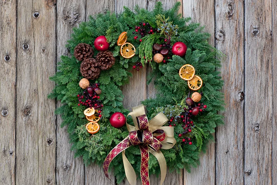 Christmas wreath with pinecones, dried orange slices, red berries, apples, and a red and gold ribbon on weathered wood.