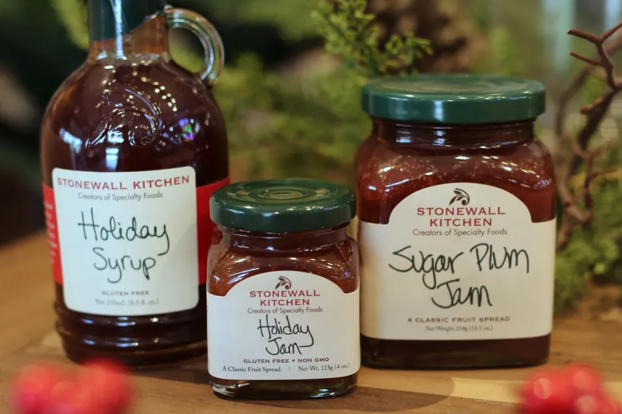 Stonewall Kitchen Holiday Syrup, Holiday Jam, and Sugar Plum Jam jars on wooden table with festive background.