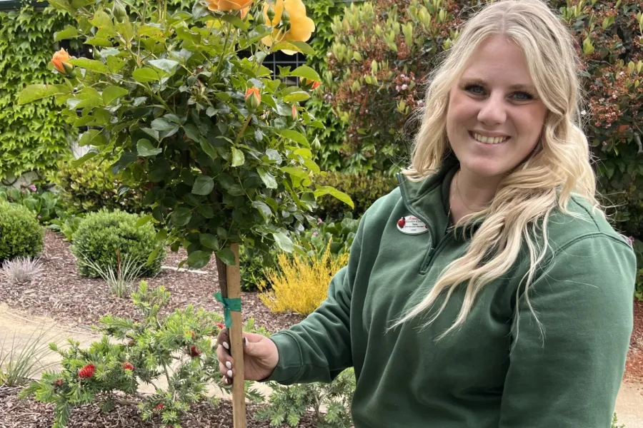 Woman in green sweater smiling while holding a potted yellow rose tree in a colorful garden.