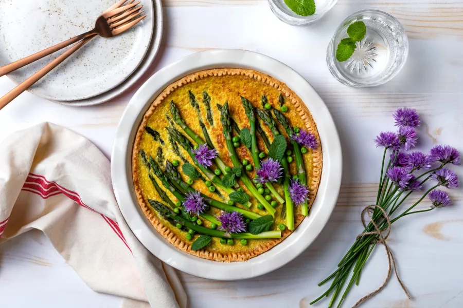 Asparagus and pea tart garnished with purple flowers and greens on a white plate with cutlery and water glasses.
