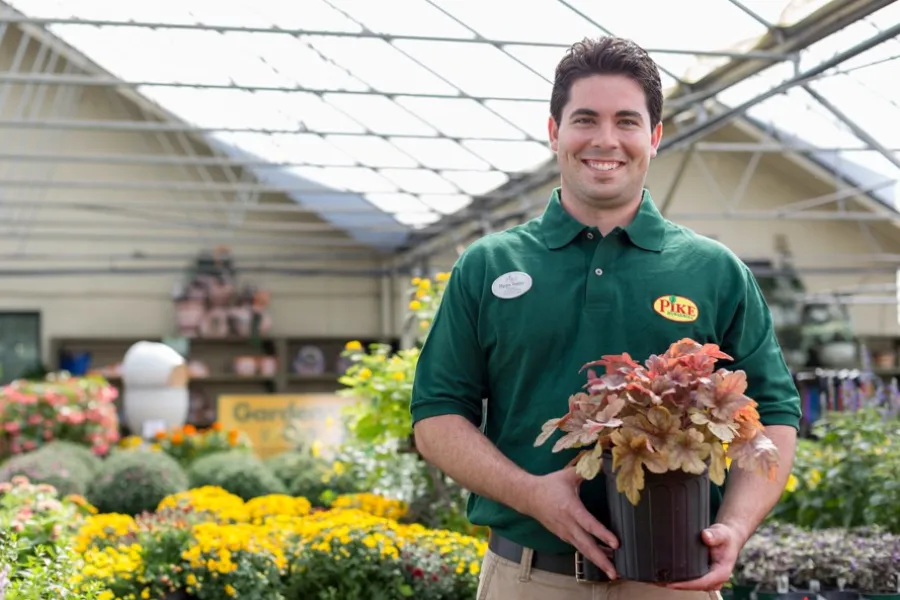 Smiling gardener in green shirt holding a potted plant inside a bright nursery with colorful flowers.