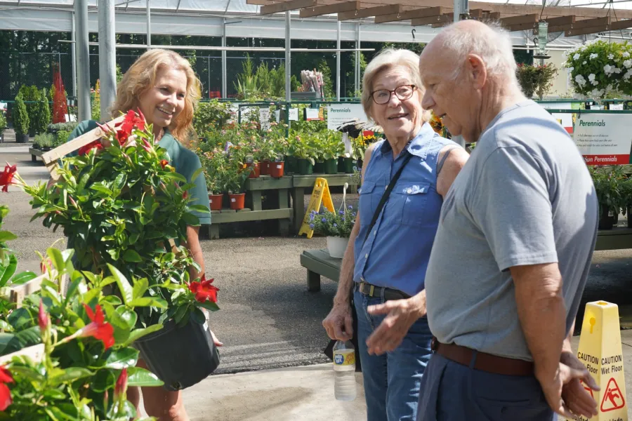 Senior couple shopping for plants with a smiling saleswoman in a bright greenhouse nursery filled with flowers.