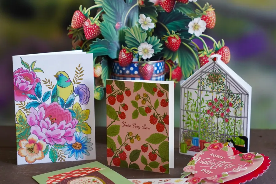 Greeting cards with floral, bird, strawberries, and Valentine designs arranged on a wooden surface with strawberry plant in background.
