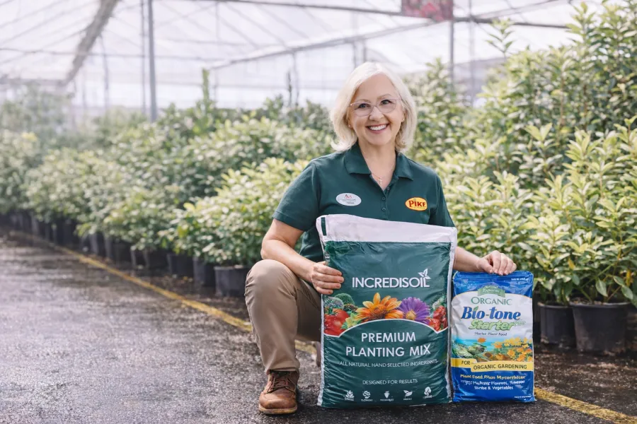Smiling woman in greenhouse holding Incredisoil premium planting mix and Organic Bio-tone gardening products.