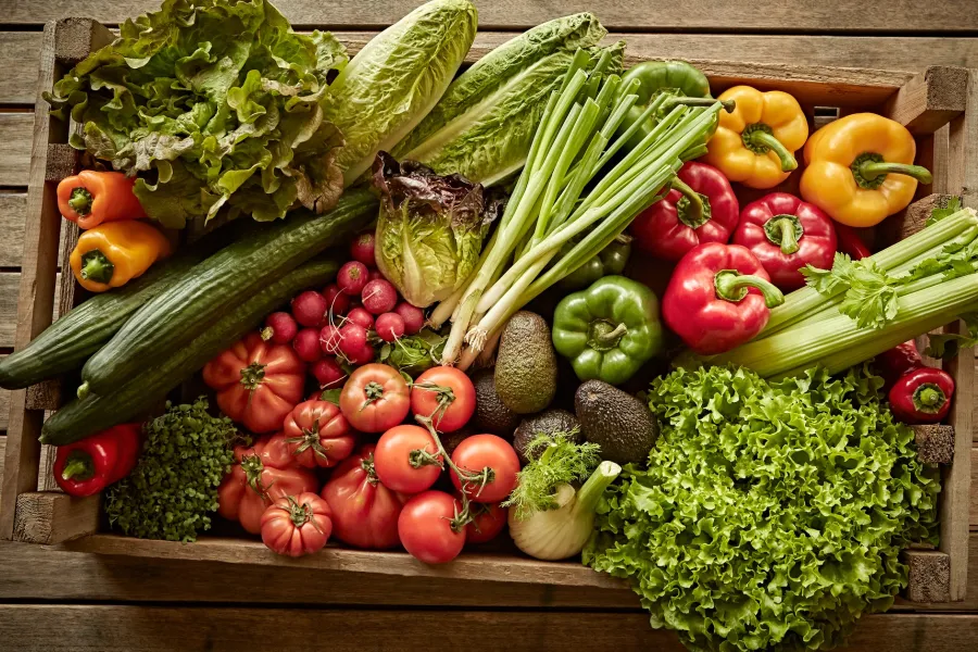 Wooden crate filled with fresh vegetables including lettuce, tomatoes, bell peppers, cucumbers, celery, and avocados.