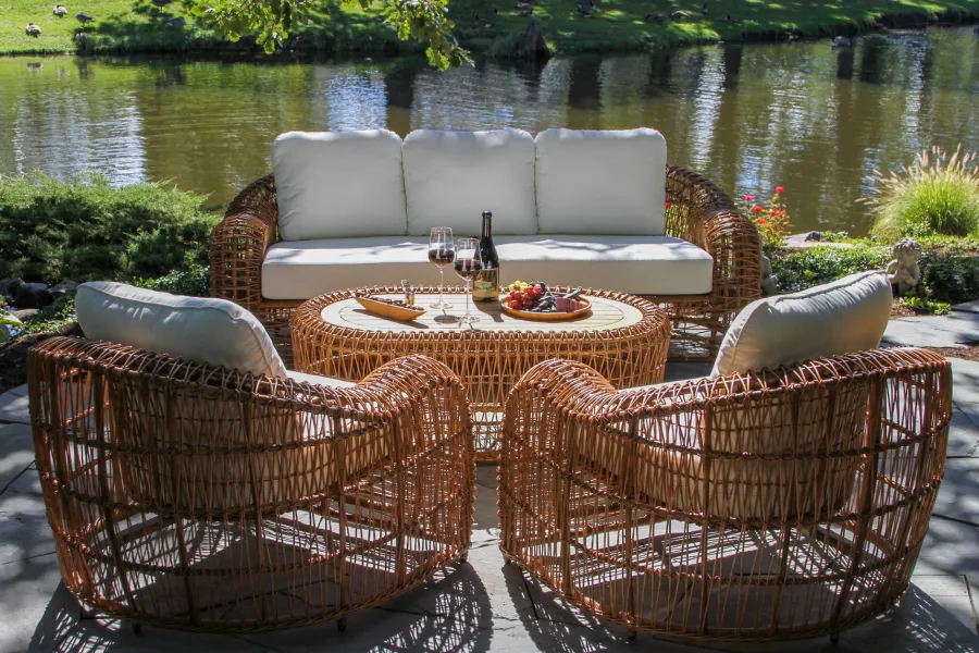 Outdoor wicker patio furniture set with white cushions by a peaceful lakeside under dappled sunlight.