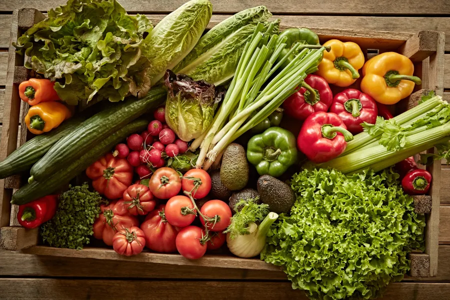 Wooden crate filled with fresh vegetables including tomatoes, peppers, cucumbers, lettuce, celery, and radishes.