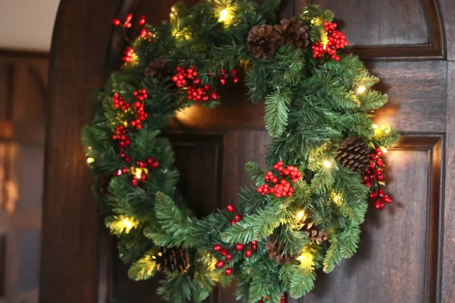 Festive Faux Christmas wreath with pinecones, red berries, and warm white lights on a wooden door.