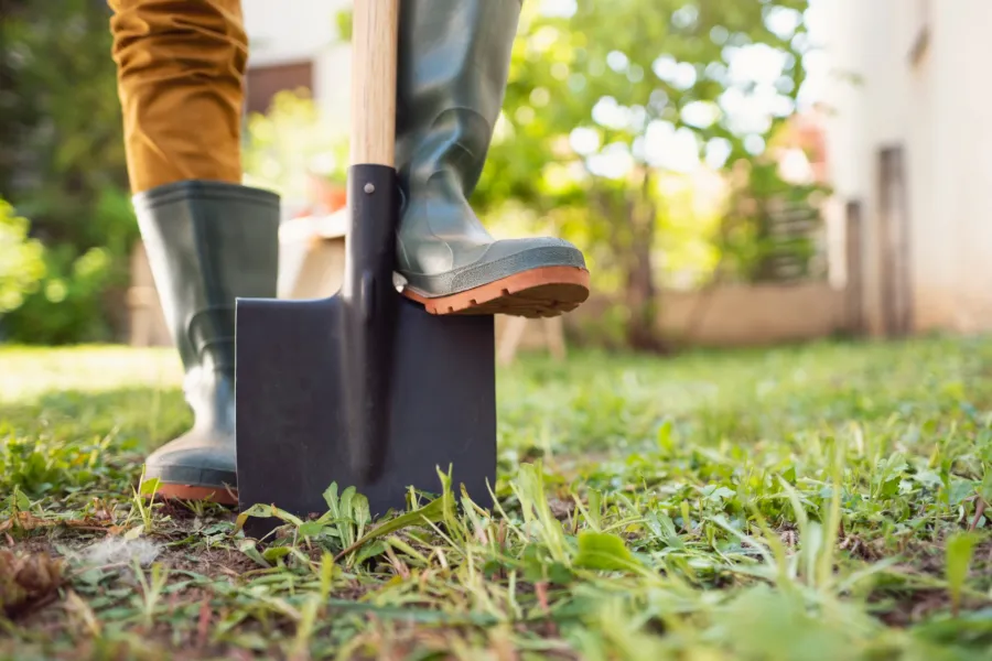 Person wearing green boots using a shovel to dig in a garden with grass and weeds on a sunny day