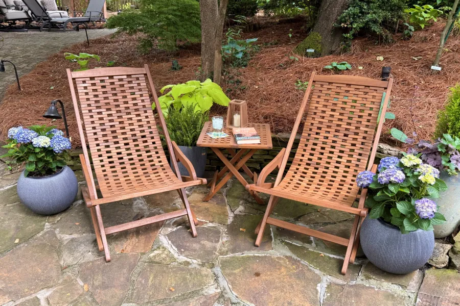 Two wooden woven lounge chairs and a small table on a stone patio surrounded by potted plants and greenery.