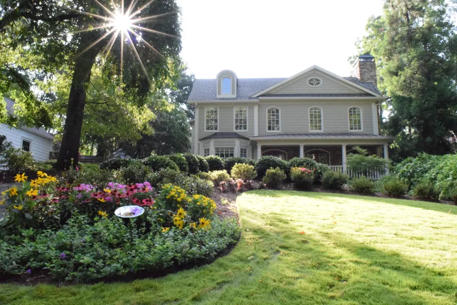 home with a landscaped front yard including lush green grass, shrubs, colorful flowers, a bird bath, and the morning sun peaking through a tree