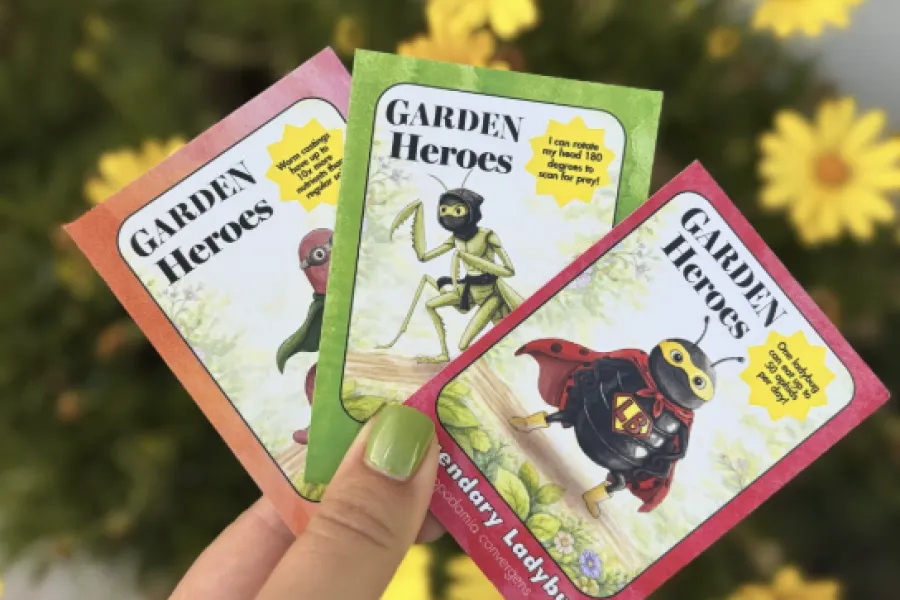 Hand holding three colorful Garden Heroes cards featuring cartoon garden insects against yellow flowers background