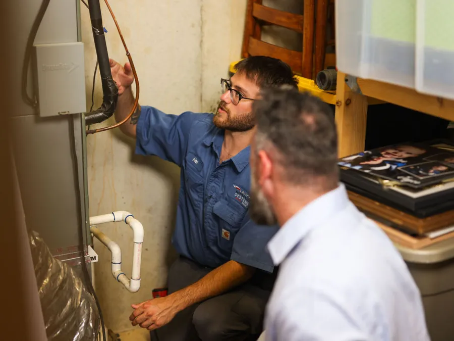 Technician in blue uniform inspecting a furnace while an older man watches closely in a basement setting.