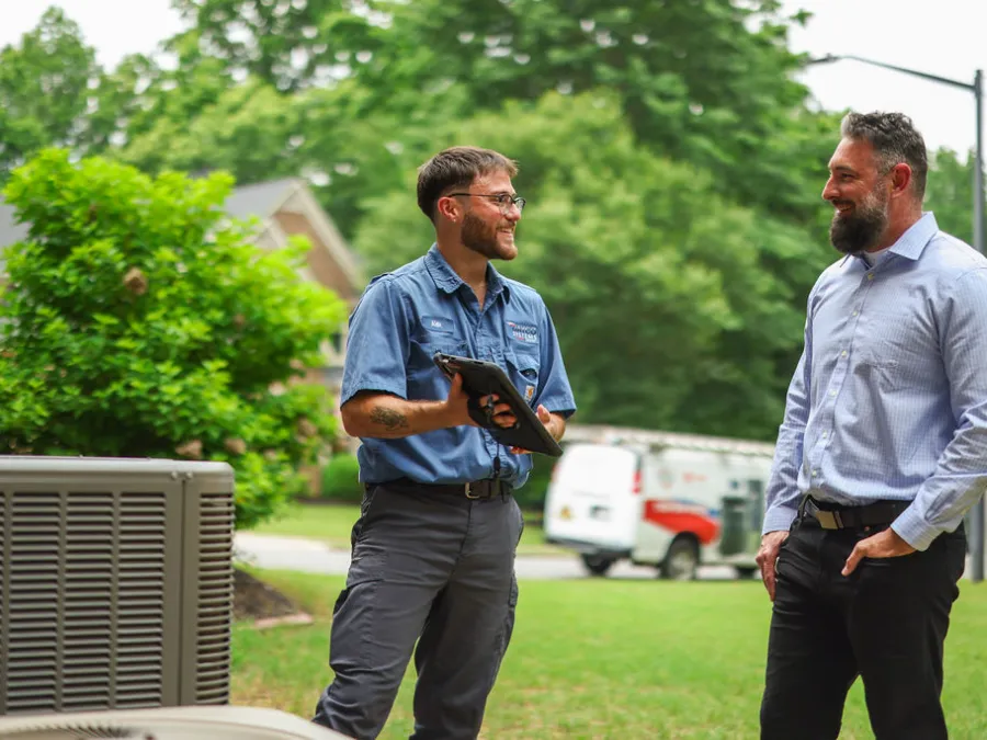HVAC technician consulting with homeowner outside near air conditioning unit and service van in a residential neighborhood.