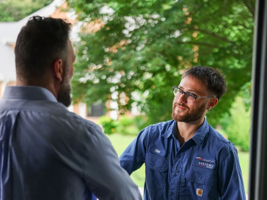 Technician in blue uniform greeting a man at a front door, holding a tablet outside a house with greenery.
