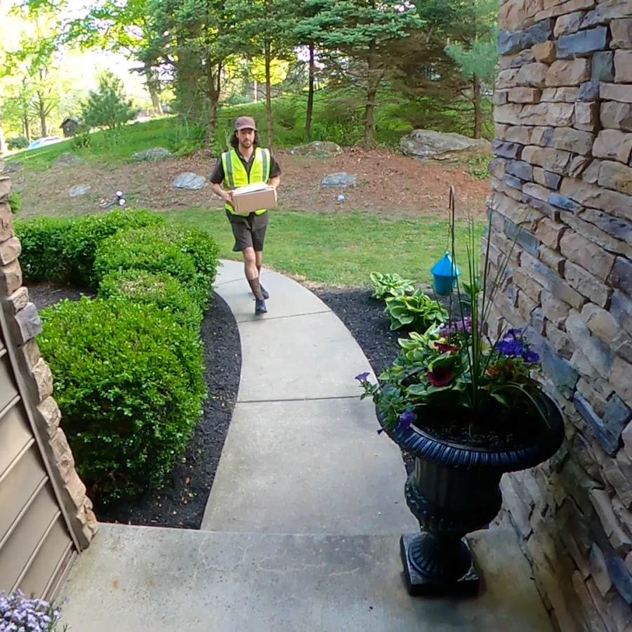 Delivery person in safety vest walking on a curved path carrying a package toward a house entrance