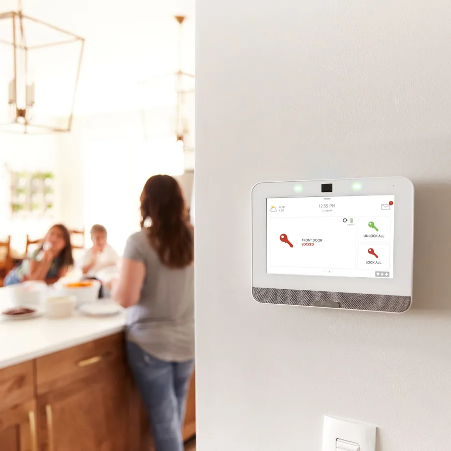 Home security system touchscreen showing locked front door with family in kitchen in background