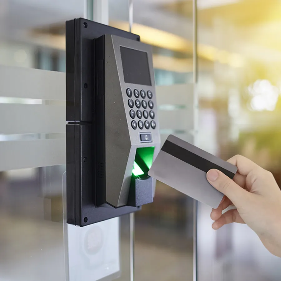 Hand holding access card near fingerprint scanner and keypad on glass door system with sunlight in background