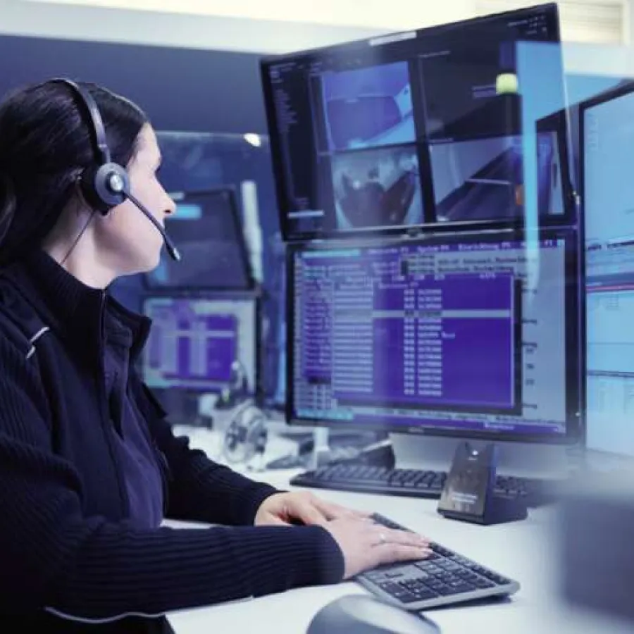 Woman with headset monitoring multiple computer screens with code and surveillance footage in control room.