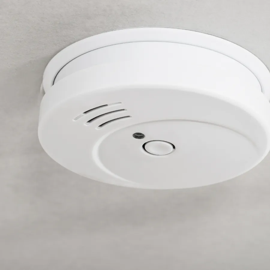 White smoke detector mounted on a ceiling with a test button and ventilation openings
