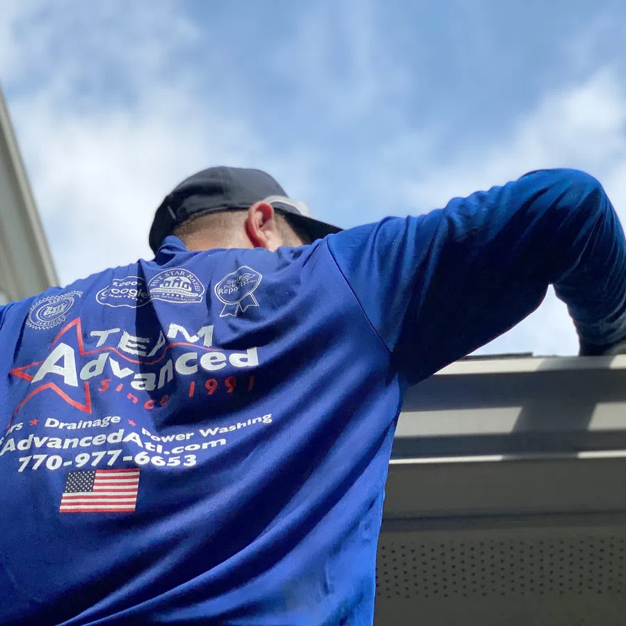 A worker in a blue shirt performing maintenance on a home's gutter under a clear sky.