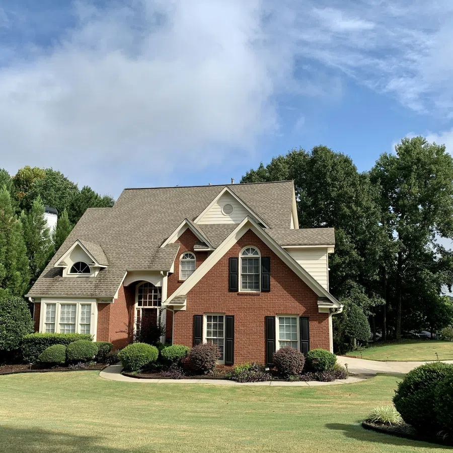 Two-story brick and beige siding house with manicured lawn and trees under a blue sky with clouds