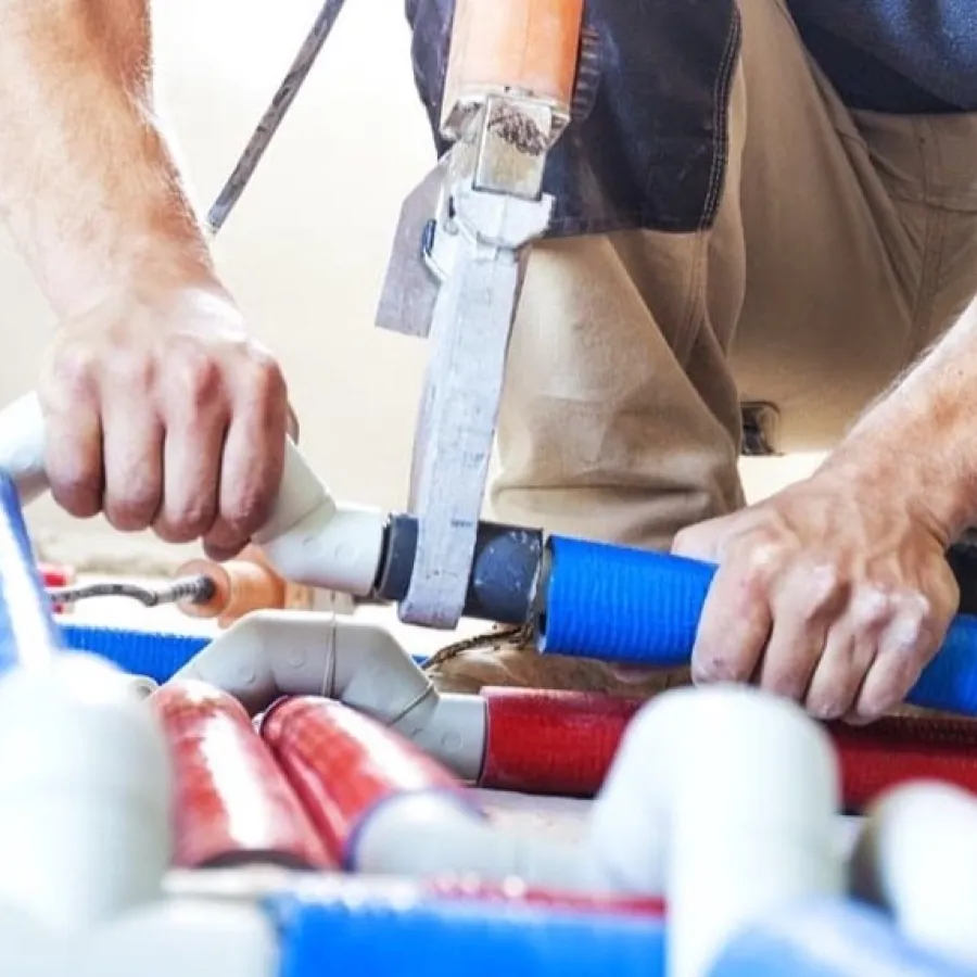 Plumber working with blue and red pipes using a cutting tool on a concrete floor in construction site.