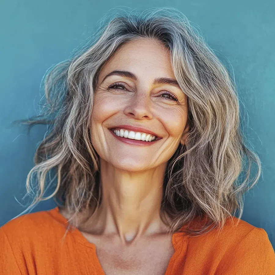 Smiling mature woman with gray wavy hair wearing an orange top against a teal background.